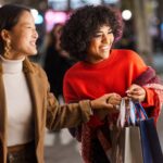 Women enjoying Black Friday holiday shopping, smiling and carrying gift bags under festive Christmas lights.