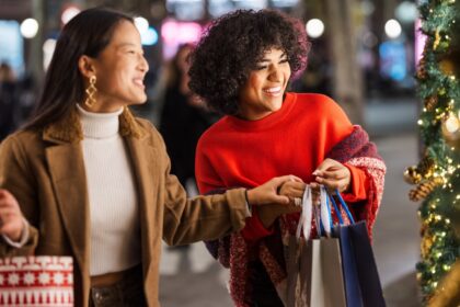Women enjoying Black Friday holiday shopping, smiling and carrying gift bags under festive Christmas lights.