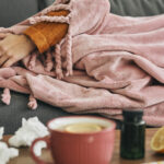Woman resting sick under a blanket with tea, tissues, lemons, and medicine on the table during cold and flu season.