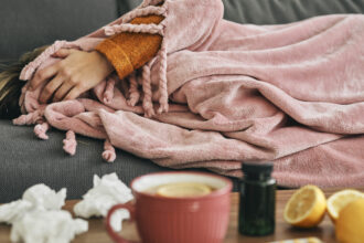 Woman resting sick under a blanket with tea, tissues, lemons, and medicine on the table during cold and flu season.