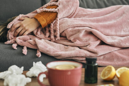 Woman resting sick under a blanket with tea, tissues, lemons, and medicine on the table during cold and flu season.